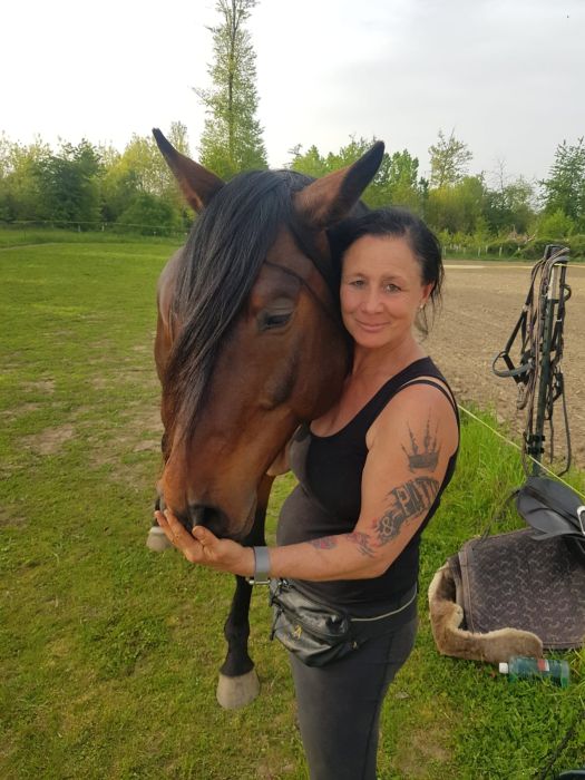Eine Frau (Manuela Rieker) steht auf einer Wiese neben einem braunen Pferd und hält dessen Kopf an ihre Wange. Sie lächelt in die Kamera und trägt ein schwarzes Top, eine Bauchtasche und hat tätowierte Arme. Im Hintergrund ist ein Reitplatz mit Bäumen zu sehen.