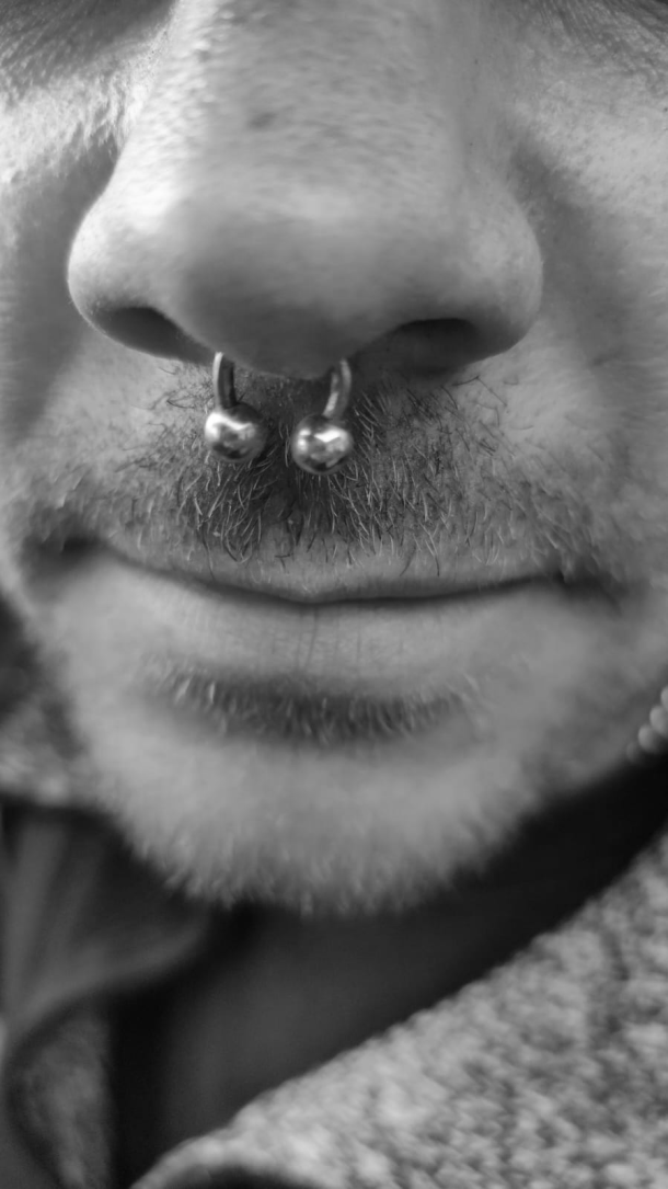 Nahaufnahme einer Nase mit einem silbernen Septum-Piercing, das zwei Kugeln an den Enden hat. Leichter Bartwuchs ist um den Mund zu erkennen.