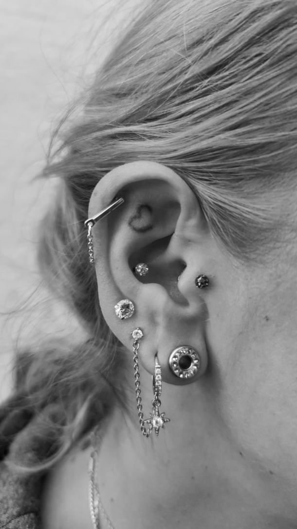 Seitenansicht eines Ohrs mit zahlreichen Ohrpiercings, darunter Stecker, Ringe, ein Industrial-Piercing sowie ein gedehntes Ohrloch. Ein kleines Herz-Tattoo befindet sich in der Ohrmuschel.